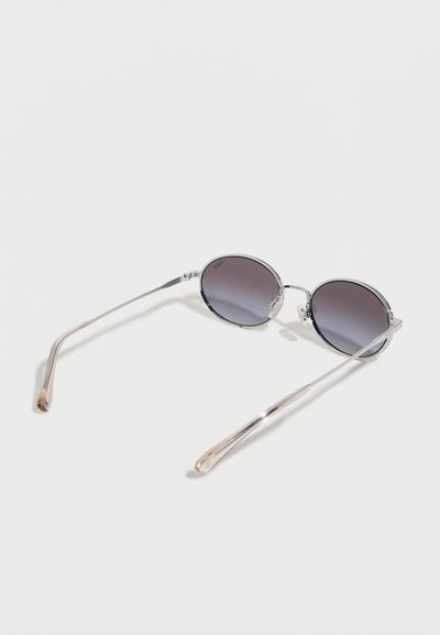 Στρογγυλά γυαλιά ηλίου με μεταλλικό σκελετό, σκούρους φακούς με χρωματισμό και διαφανή μπράτσα σε απλό ανοιχτό φόντο.
