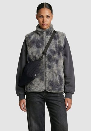 Femme portant un gilet zippé duveteux gris et noir sur un sweat-shirt foncé, avec un sac bandoulière noir, debout devant un fond clair uni.