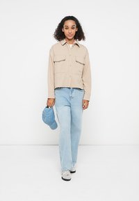 Camicia beige a maniche lunghe con due tasche sul petto, abbinata a jeans a gamba larga di colore blu chiaro. Un berretto blu è tenuto nella mano destra del soggetto.