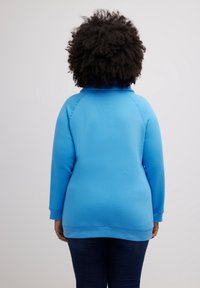 Sudadera azul con textura lisa, mangas raglán y cuello alto. Tiene un ajuste holgado y está combinada con vaqueros oscuros.
