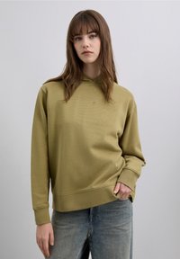 Olivgrön huvtröja med långa ärmar, ribbstickade muddar och en avslappnad passform. Har en sydd logga på bröstet och en känguruficka.