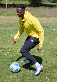Gul hættebluse, sorte bukser og lilla fodboldstøvler. Spilleren dribler en hvid og blå fodbold på græs.