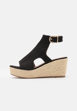 Black wedge sandal made of suede with an open toe, cutout design, and ankle strap, featuring a textured, woven jute platform heel.