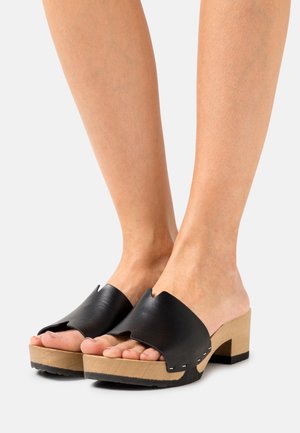 Des pieds portant des sabots ouverts en cuir noir avec des talons en bois clair sur un fond blanc.