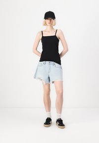 Czarny top na ramiączkach, jasnoniebieskie poszarpane szorty dżinsowe, czapka w kolorze czarnym, czarne sneakersy z beżową podeszwą, białe skarpetki do kostek. Neutralne tło.
