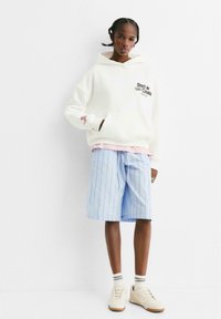 Personne portant un sweat à capuche blanc avec le texte "Start Create", un short bleu à rayures de longueur genou, des chaussettes blanches et des baskets beiges, se tenant contre un mur blanc.