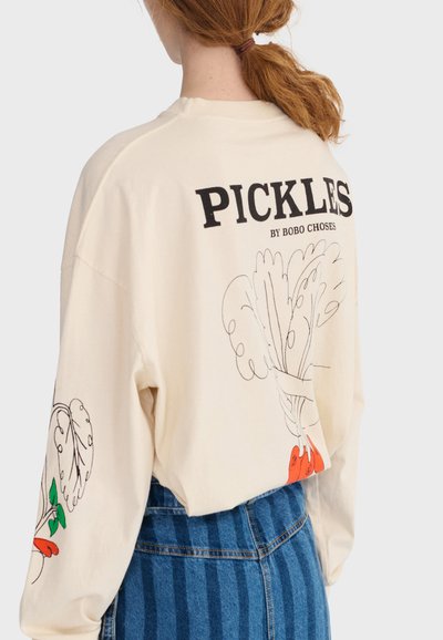 Osoba s crvenom kosom u repu nosi kremasti duks s natpisom "PICKLES BY BOBO CHOSES" i ilustracijama povrća na leđima i rukavu.