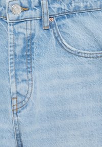 Jean en denim bleu clair présentant une texture lisse, une décoloration subtile, des coutures en cuivre, et une poche avant. Détail de fermeture par bouton en métal visible.