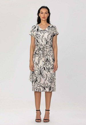 Femme debout, vêtue d'une robe blanche mi-longue avec des motifs floraux abstraits noirs et des talons noirs à bride cheville, sur un fond clair uni.