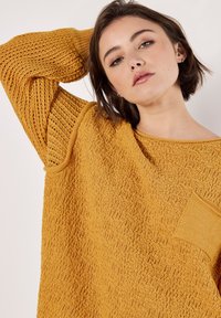 Senfgelbes Strickpullover mit strukturiertem Stoff, weiten Ärmeln und einer kleinen Fronttasche. Lockere Passform mit rundem Ausschnitt und gerippten Abschlüssen.