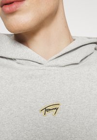 Sweatshirt gris à capuche avec une texture douce, arborant un logo "Tommy" brodé en noir et or sur la poitrine.