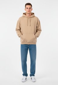 Ung man står med händerna i fickorna, iklädd en beige hoodie, blåjeans och vita sneakers med svarta ränder mot en vit bakgrund.