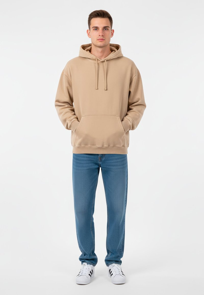 Ung man står med händerna i fickorna, iklädd en beige hoodie, blåjeans och vita sneakers med svarta ränder mot en vit bakgrund.