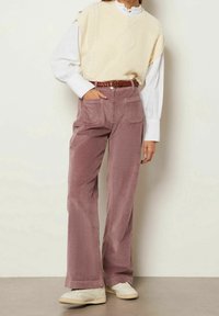 Beige stickad väst över en vit skjorta, i kombination med mjuka mauve kordbyxor. Flätat brunt bälte, texturerad tyg, och krämfärgade sneakers.