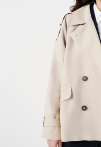 Trench coat beige con bottoni doppi, ampio colletto e spalline. Tessuto morbido con una vestibilità leggermente strutturata.