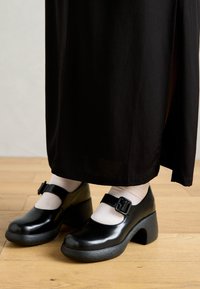 Black patent leather Mary Jane shoes with a rounded toe, a buckle strap, and a chunky heel. Worn with light-colored ankle socks.
