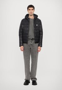 Chaqueta acolchada negra con capucha, que cuenta con un cierre frontal y un parche con el logo. Lleva puesta sobre una camiseta gris y unos jeans grises, combinada con zapatos negros.