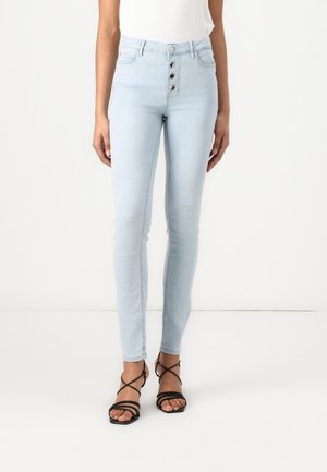 Mujer vistiendo jeans ajustados de tiro alto en azul claro con cuatro botones delanteros y sandalias negras de tacón con tiras.