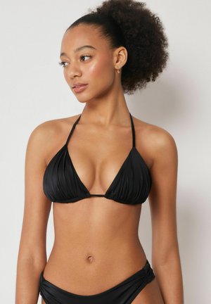 Junge Frau mit lockigem Haar, die einen schwarzen Neckholder-Bikinibadeanzug und passende Unterteile trägt und vor einem schlichten hellen Hintergrund steht.