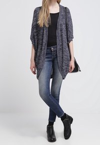 Donna con un top nero, un cardigan grigio a righe, jeans blu, stivali neri alla caviglia con borchie e una borsa a tracolla nera.