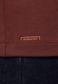T-paita, ruskeanpunainen, jossa on ribattu pinta ja pieni logo-etiketti "9STRAATJES" oranssina helmassa. Taustalla näkyy denimi.