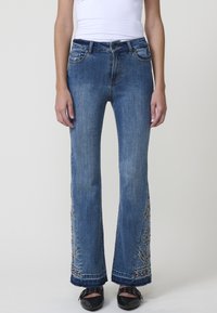 Jeans de mezclilla azul acampanados con patrones florales bordados en el dobladillo, cinco bolsillos y cierre de botón. La tela tiene una textura desgastada.