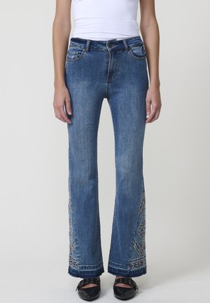 Jeans de mezclilla azul acampanados con patrones florales bordados en el dobladillo, cinco bolsillos y cierre de botón. La tela tiene una textura desgastada.