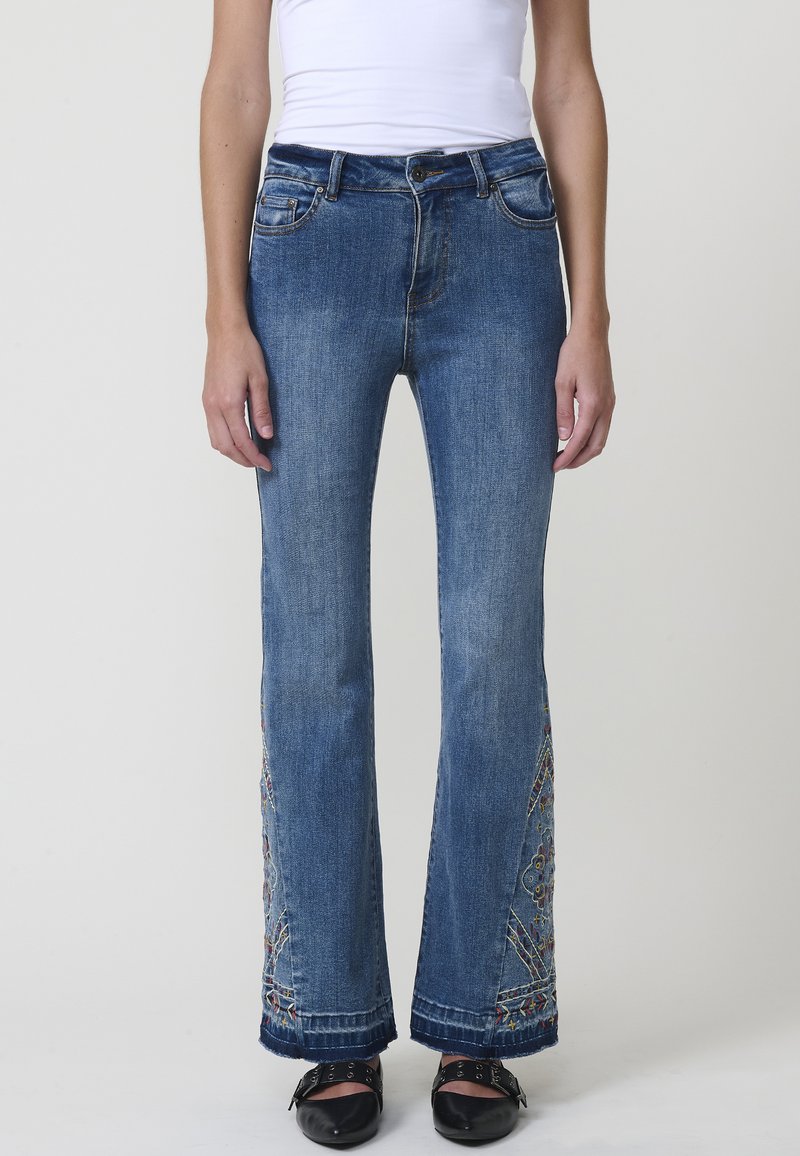 Jeans de mezclilla azul acampanados con patrones florales bordados en el dobladillo, cinco bolsillos y cierre de botón. La tela tiene una textura desgastada.