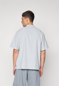 Light gray short-sleeve shirt with a collar, featuring a textured finish and a relaxed fit. The hem is straight with a small branded tag.