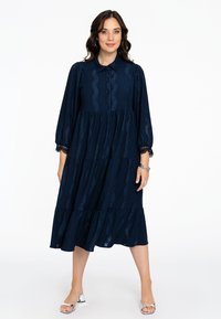 Marineblaue Stufenkleid mit langen Ärmeln, Kragen, Knopfverschluss und Spitzenmuster-Details. Kombiniert mit silbernen Absatzsandalen für einen Kontrast.