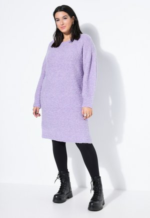 Kvinde står iført en lang, lavendelfarvet, pelsagtig sweaterkjole, sorte leggings og sorte snørestøvler mod en hvid baggrund.