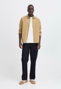 Beige button-up shirt over a white t-shirt, worn with black cargo pants and grey sneakers, featuring two chest pockets and a relaxed fit.
