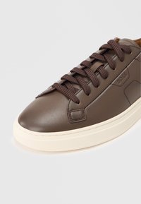 Brown leather sneaker with dark brown laces, cream outsole, and subtle stitched details. Features logo on the side panel.