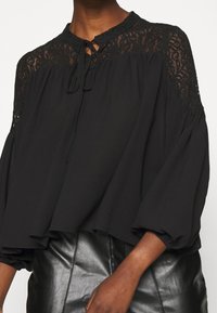 Blouse noire avec détails en dentelle sur les épaules et un col montant. Comprend un cordon de serrage et des manches amples et bouffantes. Tissu lisse.