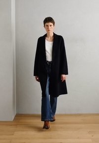 Manteau long noir sur une chemise blanche, associé à un jean bleu évasé et des chaussures brunes. Comprend une fermeture à un bouton et des manches longues.