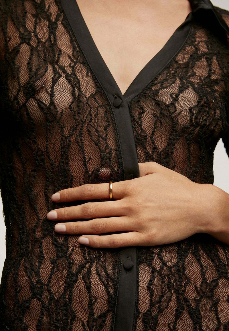 Prenda de encaje negra con intrincados patrones florales, que presenta un frente con botones y un cuello de satén. Anillo dorado en una mano colocada sobre la tela.