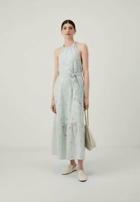 Ljusblå, ärmlös maxiklänning med halterneck, knytmidja, spetsliknande mönster och lagerkjol. Tillbehör inkluderar en beige väska och slip-on skor.