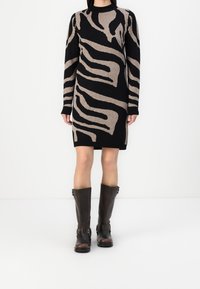 Femme portant une robe-pull à manches longues noire et beige avec un motif abstrait, et des bottes en cuir marron foncé montant jusqu'au genou.