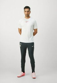Camisola branca de manga curta para atletismo com logótipo da Nike vermelho, combinada com calças de treino Nike verde-escuro e chuteiras de futebol cor-de-rosa. Design minimalista.