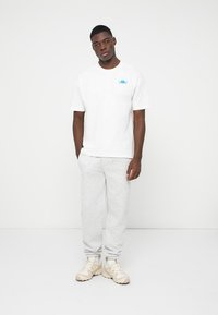 Tricou alb din bumbac cu un mic logo albastru, asociat cu pantaloni de trening gri deschis și adidași bej, stând în fața unui fundal simplu.
