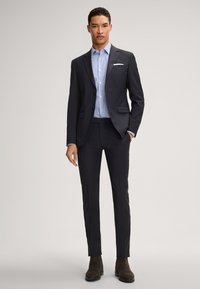 Completo blu navy con una vestibilità tailored, caratterizzato da due bottoni e rever a punta, abbinato a una camicia a righe azzurra e stivali in suede marroni.