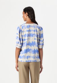 Kottongbluse med blå og beige tie-dye mønster, rund hals, og korte, puffete ermer. Kombinert med beige bukser.