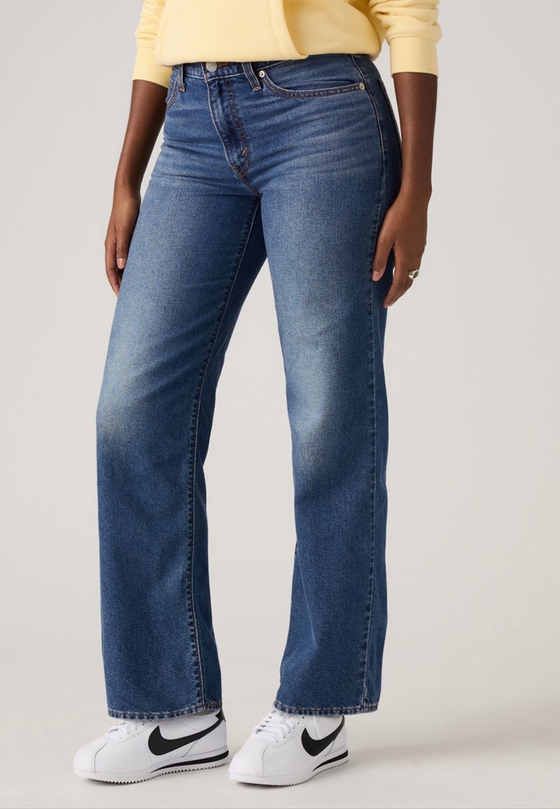 Personne portant un jean bleu coupe droite, des baskets blanches Nike avec un swoosh noir et un haut à manches longues jaune pâle.