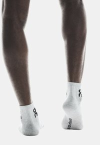 Calcetines deportivos blancos con puños acanalados y detalle del logo en negro, descansando sobre los pies de una persona con piernas musculosas sobre un fondo liso.