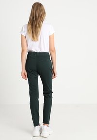 Pantaloni slim-fit verde scuro realizzati in una morbida stoffa, con vita media, tasche posteriori, indossati con una maglietta bianca semplice e scarpe da ginnastica.
