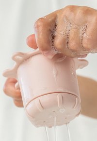 Roze siliconen beker die wordt ingedrukt, waarbij water en zeepbellen eruit stromen. Heeft een gestructureerde grip en een uitwaaierende rand.