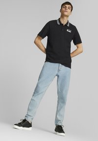 Svart polo skjorte med hvite striper ved kragen, logo detaljer på venstre bryst, kombinert med lyseblå jeans og svarte sneakers.