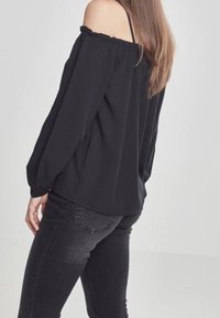 Mustassa olkapäiltä paljastavassa pitkähihkaisessa puserossa on röyhelökaulus, rento malli ja jotain valkoisia hihoja, yhdistettynä tummiin farkkuihin.