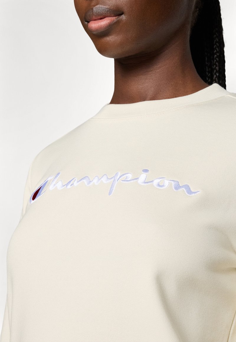 Cremfarbener Sweatshirt mit Rundhalsausschnitt und gesticktem "Champion"-Logo in Blau und Rot. Weicher Stoff, sichtbare Nähte.