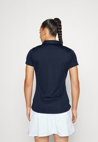 Sötétkék, rövid ujjú póló, gallérral, sima textúrával és testhezálló fazonnal. A hátul ívelt alsó szegéllyel rendelkezik, amely a csípőnél végződik.